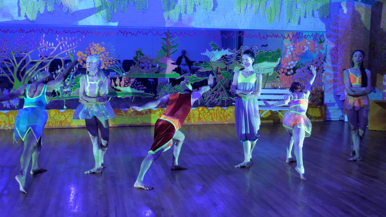 Five dancers - three (two men and a woman) facing upstage in a deep horizontal lunge with right arm extended while two woman dancers stand between them left leg tucked behind. All are costumed in exuberant colors and styles - little mesh tutu, longer sheer skirt, zigzag bottomed skirt, a wrap skirt. The tops are body hugging and outlined in fluorescent green. The back drop is filled with painted flowers and trees.